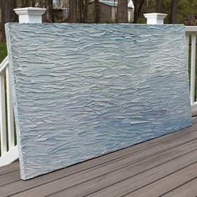A abstract painting with blue textured brushstrokes is seen at an angle on a deck at the artists studio.