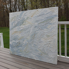 An abstract painting with thickly textured blueish, orangish and greenish beige brushstrokes is seen at an angle on the artists deck outside.