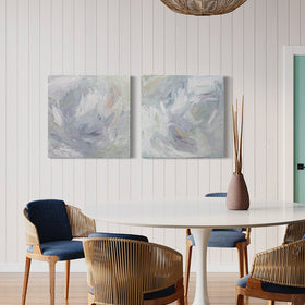 A pair of thickly painted paintings in teal, sea foam green, celadon, white, lavender and yellow by Teodora Guererra hang on a white wall in a kitchen. A white pedestal table surrounded by modern wicker type chairs with blue cushions sits in front of the paintings with a ceramic vase and sticks sitting on the table. Over the table hangs a wicker light and a door to the right can be seen in apple green.