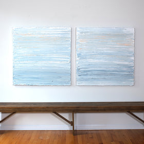 An pair of paintings with thick impasto brushstrokes of blue, white, teal and hints of orange above a bench.