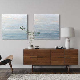 An pair of paintings with thick impasto brushstrokes of blue, white, teal and hints of orange above a wood credenza with a plant sitting on the credenza in front of the paintings. A chair sits to the left side.