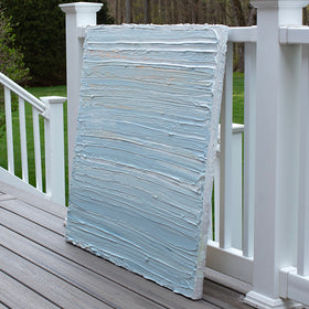 An side view of an abstract painting with thick impasto brushstrokes of blue, white, teal and hints of orange paint sits on a deck at the artists studio.