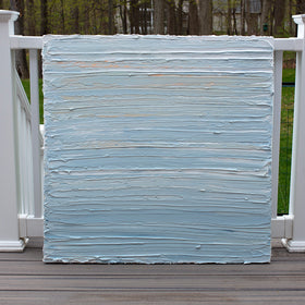 An abstract painting with thick impasto brushstrokes of blue, white, teal and hints of orange paint sits on a deck at the artists studio.
