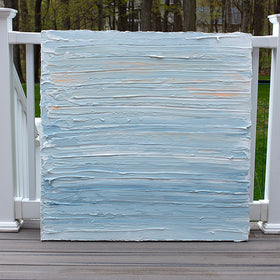An abstract painting with thick impasto brushstrokes of blue, white, teal and hints of orange paint sits on a deck at the artists studio.