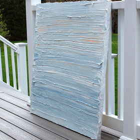 An side view of an abstract painting with thick impasto brushstrokes of blue, white, teal and hints of orange paint sits on a deck at the artists studio.