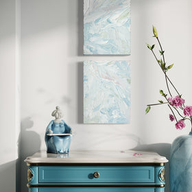 A pair of thickly painted paintings in teal, sea foam green, celadon, white and hints of rose by Teodora Guererra hang on a white wall over an ornate teal end table with white marble top. To the right is a marble vase with pink flowers and sitting on the table is a figurine of an asian woman in a teal dress. The two painting are like wall sculptures.