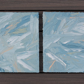 A pair of thickly painted paintings in teal, sea foam green, celadon, white and hints of orange and yellow by Teodora Guererra laying flat on a deck outside. The two painting are like wall sculptures.