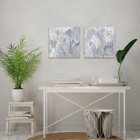 2 paintings hung as a pair shown hanging on the wall over a white table with a plant to the side. The surface of the paintings are decorated with white, blue and grey paint in a thick impasto. Wired and ready to hang.