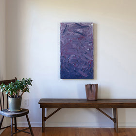 A tonal purple and pink textured abstract painting by Teodora Guererra hangs on a white wall above a wood bench with a wood tabletop sculpture and a chair with a potted plant.