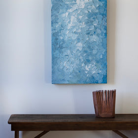 A blue and white abstract painting by Teodora Guererra hangs vertically on a white wall above a dark wood bench with a ceramic sculpture accent.